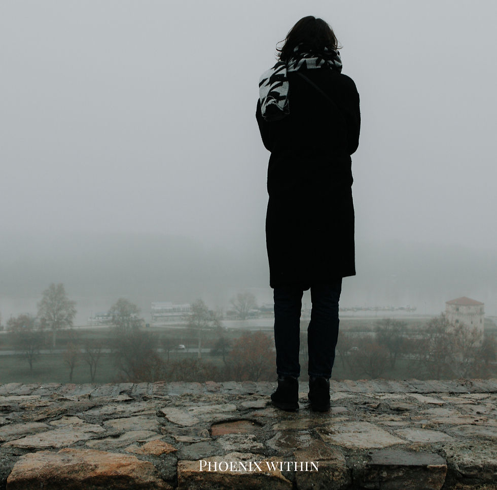 A person stands on a foggy stone wall overlooking a river. They wear a coat and scarf. Mood: contemplative.