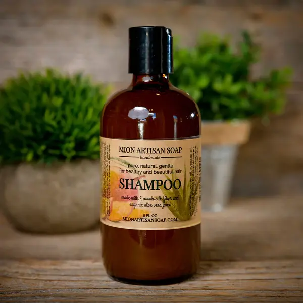 A bottle of Tussah Silk & Aloe Vera Shampoo sits on a wooden shelf with microgreens in the background.