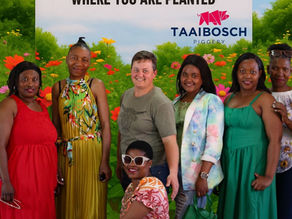 Sign reading 'Women's day 2025, bloom where you are planted'. Group of Taaibosch female employees standing in front of sign