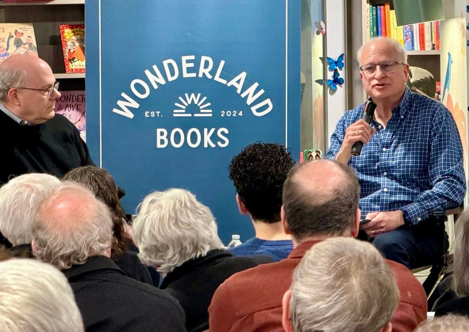 The author speaks into a microphone to a seated crowd in front of a sign that reads "Wonderland Books"