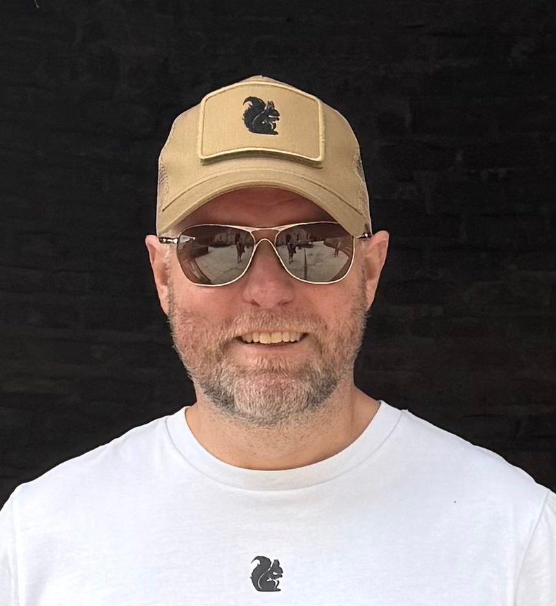 Man wearing a sand coloured trucker cap with a black squirrel logo on the front