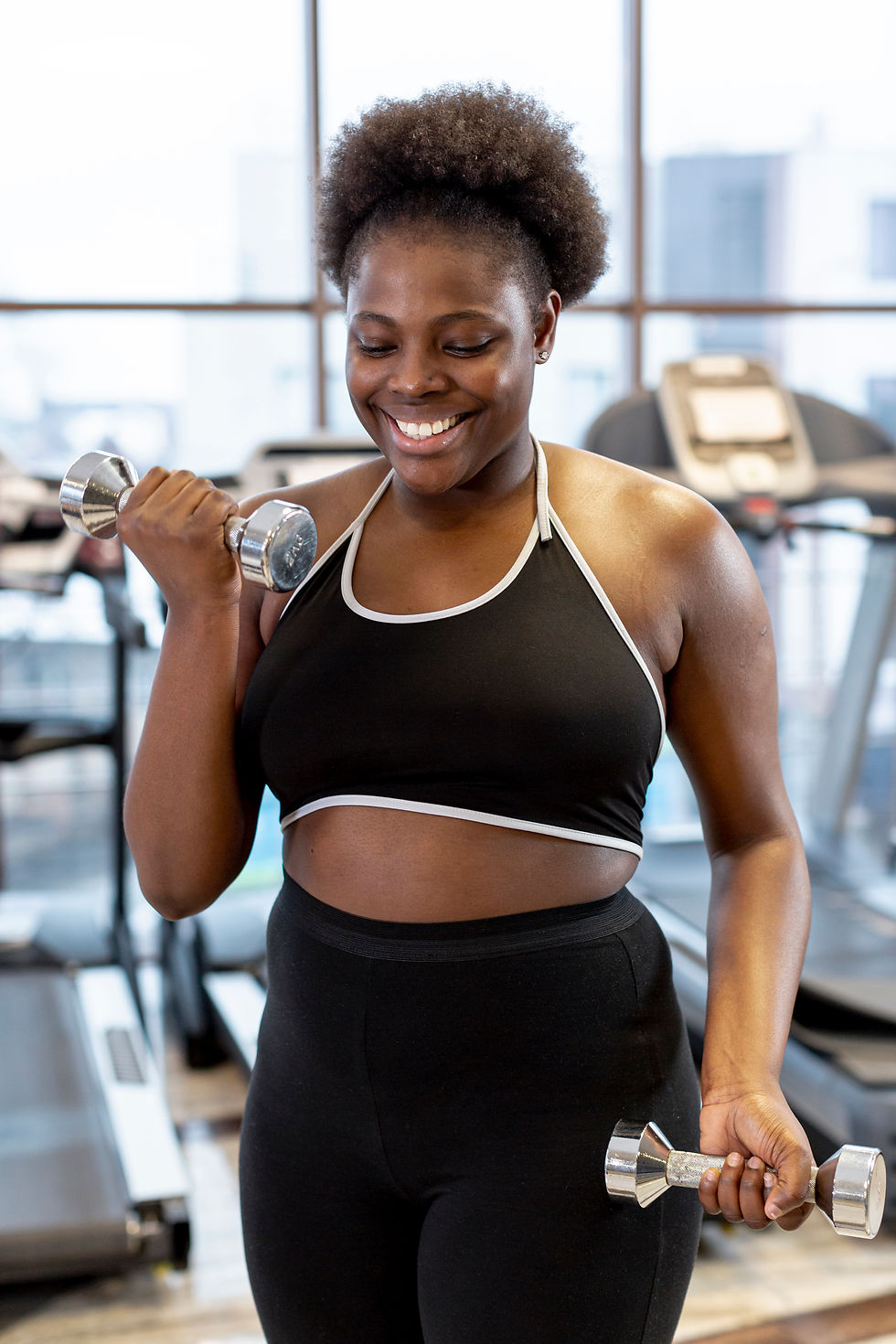 Smiling person lifting dumbbells in a gym, wearing black workout attire. Background includes treadmills and large windows.