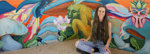 Artist Kristina Greenwood sitting on the floor, engaged with a bright and colorful mural that features giant mushrooms and fantastical creatures. 
