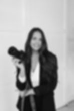 Destination wedding photographer in a black jacket holding a camera, smiling in a black-and-white photo. Bright background, relaxed and confident mood.