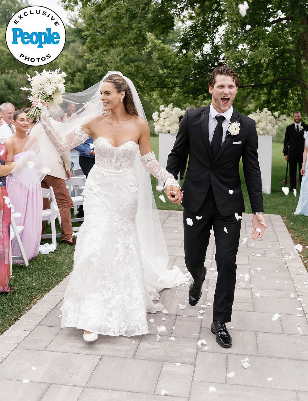 Bride in lace dress and groom in black suit joyfully exit outdoor wedding, holding hands amid white flower petals. Lush greenery surrounds them.
