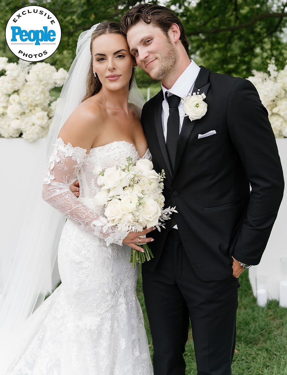 Bride in lace gown and groom in black suit stand together, smiling, outdoors with white flowers and green trees. "People Exclusive Photos" logo.