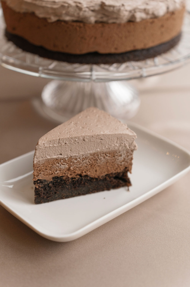 Slice of layered chocolate mousse cake on a white plate, with whole cake on a stand in the blurred background. Neutral table setting.