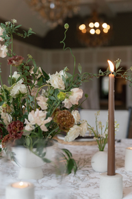 Elegant table setting with floral centerpiece, candles, and silverware on a patterned tablecloth. Chandeliers hang in the upscale venue.