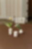 White vases with a yellow tulip and small white flowers on a brown table. A lit tealight candle nearby. Beige wall and a chair in the background.