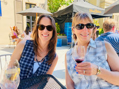 Smiling women in sunglasses toast wine at an outdoor patio under umbrellas, with Chateau St. Michelle tent behind them.