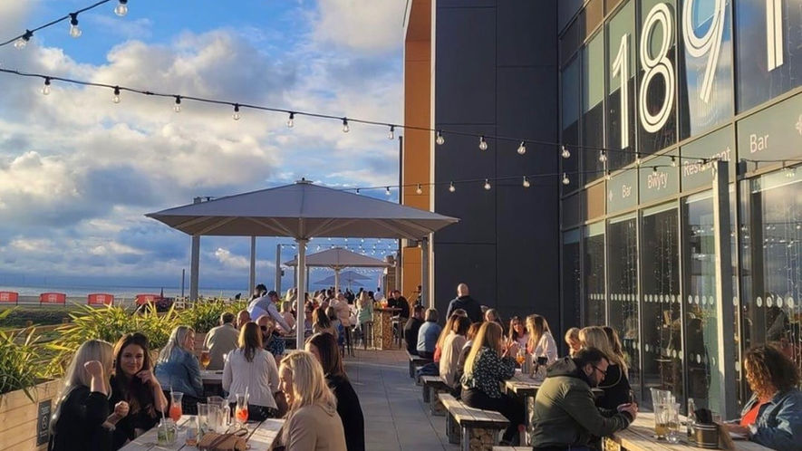 A coastal bar terrace with rustic gabion furniture, festoon lighting and large beige umbrellas in background.