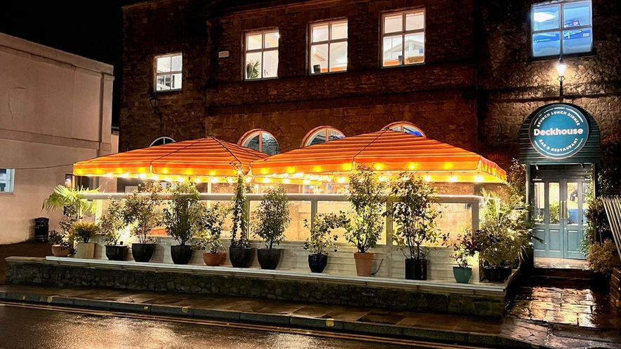 A cafe terrace at night with two orange striped jumbo umbrellas with festoon lighting.