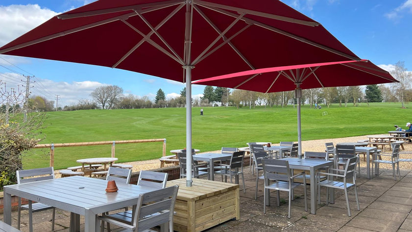 A golf clubhouse terrace with metal dining furniture and two red umbrellas on freestanding base.
