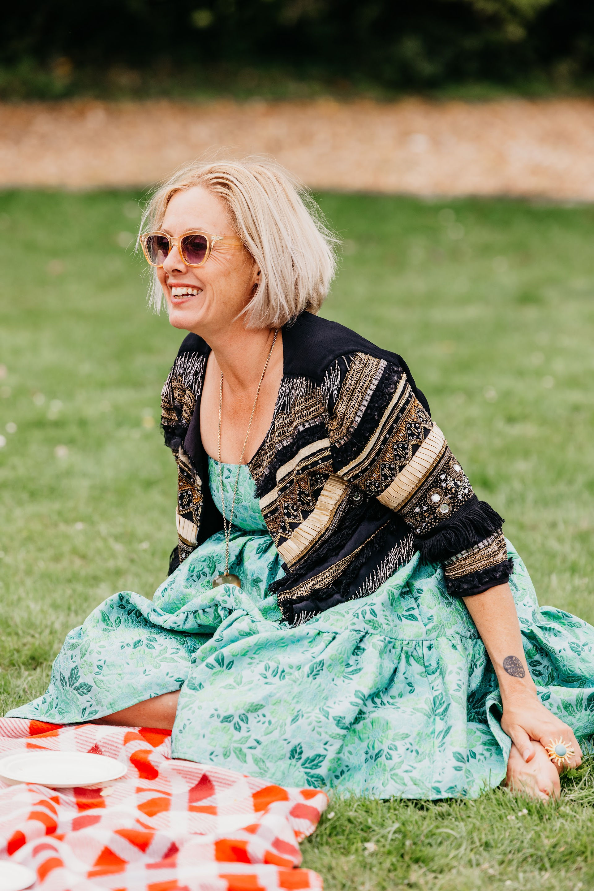 Laughing guest in sunglasses and a green floral dress sitting on an orange checked picnic rug on the lawn