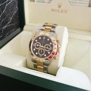 a stainless steel and yellow gold rolex daytona 16523 in a rolex box featuring a black diamond set dial, yellow gold bezel, and two tone oyster bracelet
