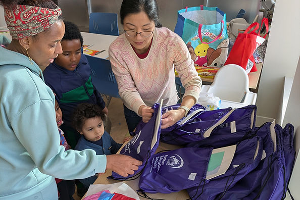 Two women and two children handed literacy kits at a table in a bright room. Colorful bags in the background.