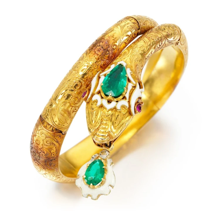 Designed as a hinged floral and foliate chased serpent, the head decorated with white enamel and set with a pear-shaped emerald, with circular-cut ruby eyes, issuing a white enamel and pear-shaped emerald drop from its mouth, inner circumference approximately 150mm.