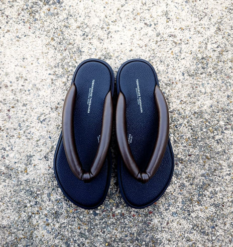 アスファルト(コンクリート)の床の上に置かれた、BEAUTIFUL SHOES(ビューティフルシューズ)の「SETTA SANDALS」。伝統的な雪駄をモダンに昇華した、ふっくらとボリュームのある黒いレザーの鼻緒とシボ感のあるインソールが特徴的なトングサンダルの写真。
