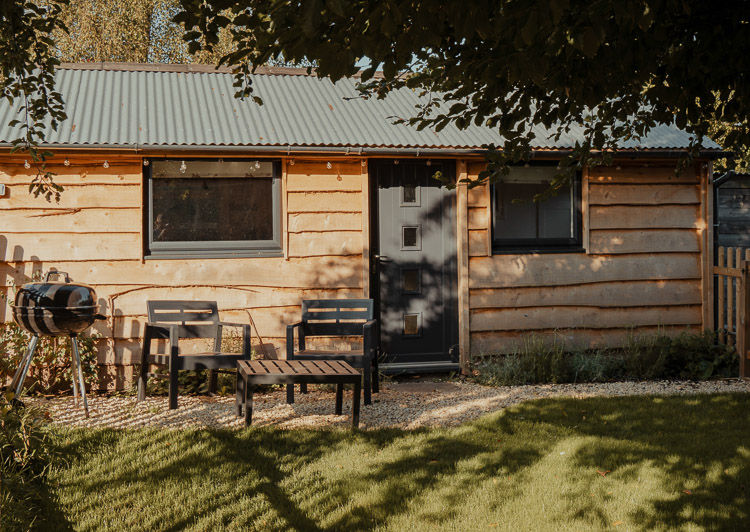 Wooden garden cabin with black chairs, table and charcoal grill in a sunny, shaded yard under trees.