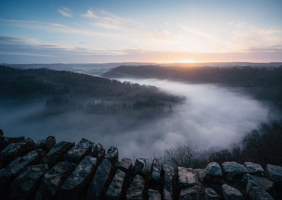 Misty valley at sunrise, viewed over a stone wall, with dark hills and soft pink-blue sky.