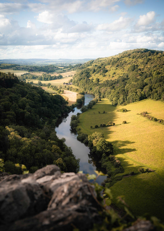 Sunlit river winds through a green valley between wooded hills under a cloudy sky, viewed from a rocky overlook.