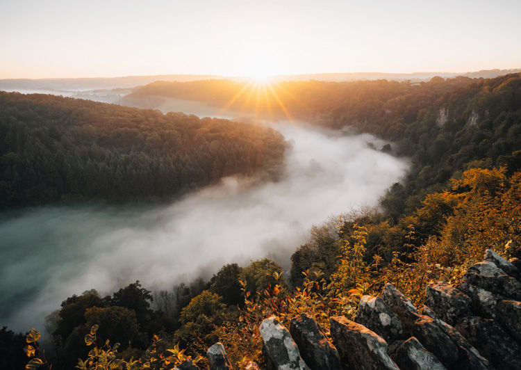 Sunrise over a fog-filled river valley with forested hills and rocky foreground.