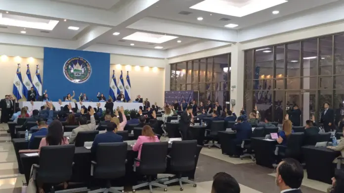 Personas en una asamblea parlamentaria votan, con banderas y escudo nacional en el fondo azul. Ambiente formal y organizado.