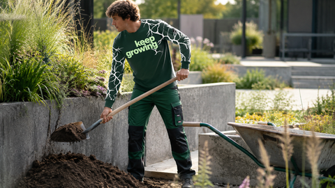 Junger Gärtner arbeitet mit einer Schaufel in einem modernen Garten und trägt ein grünes „keep growing“-Shirt.