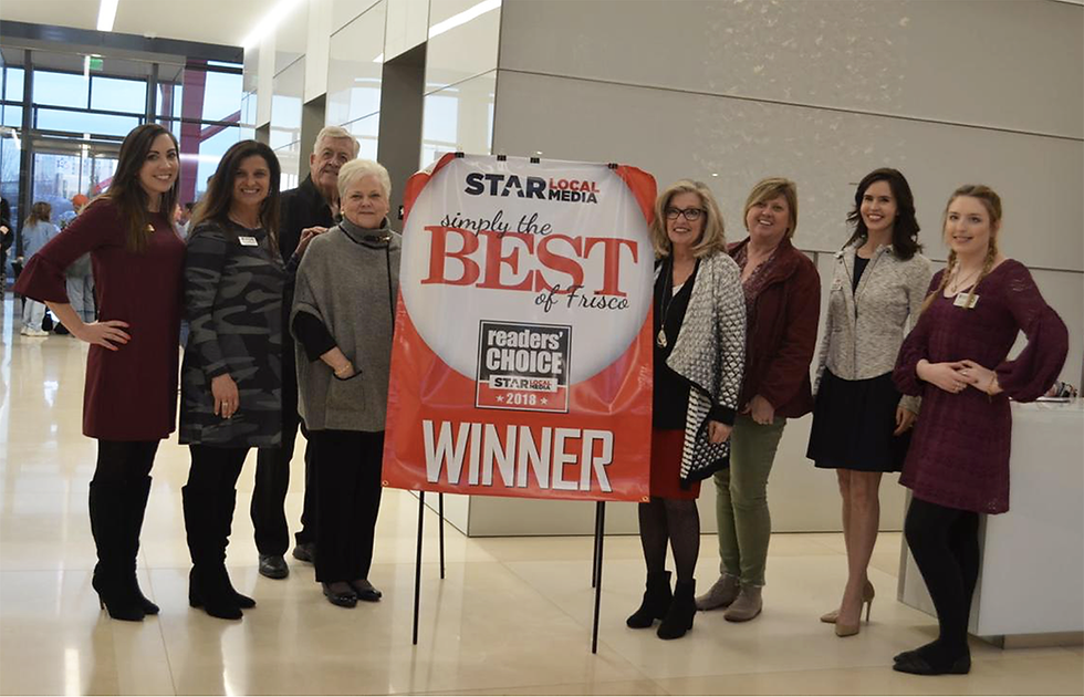 Group of eight people smiling beside a red "Best of Frisco" winner banner in a modern building. Bright setting with reflective floors.