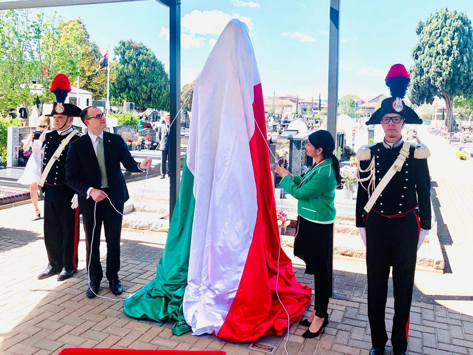 People and one woman standing besides a covered statue draped in the Italian flag during an outdoor ceremony.