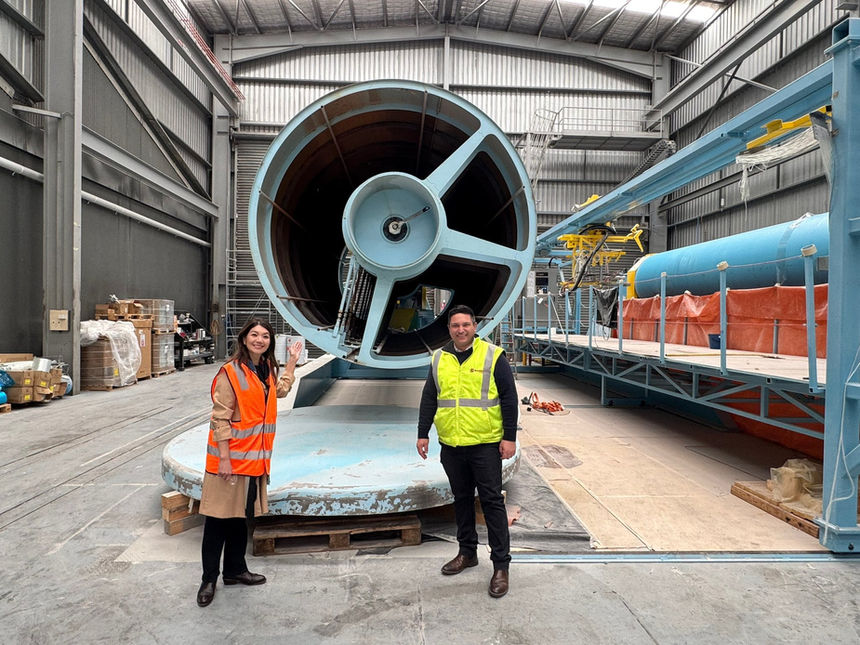Two people standing inside a manufacturing facility during a site visit, positioned in front of a large cylindrical industrial machine.