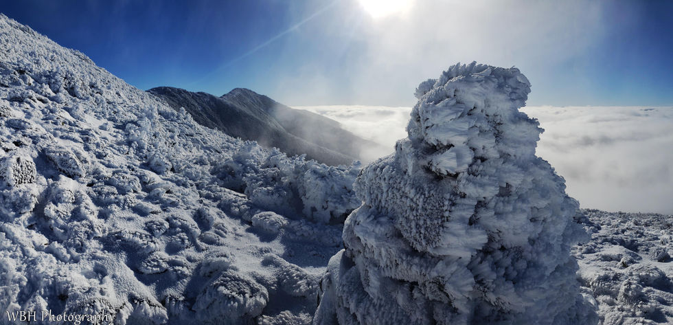 Snow-covered mountain landscape with icy formations and a cloud-filled valley under a bright sun.