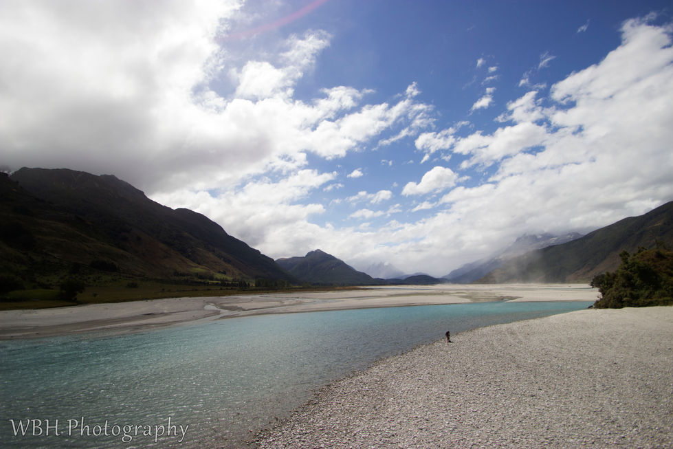 Very wide and shallow creek with rocky shore and small rocky islands with mountains on the side and blue skies and clouds in New Zealand.