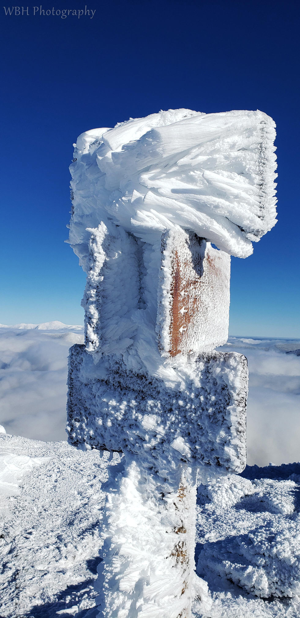 Ice-covered trail sign on a snowy mountain peak with a deep blue sky and cloud-covered landscape in the background.