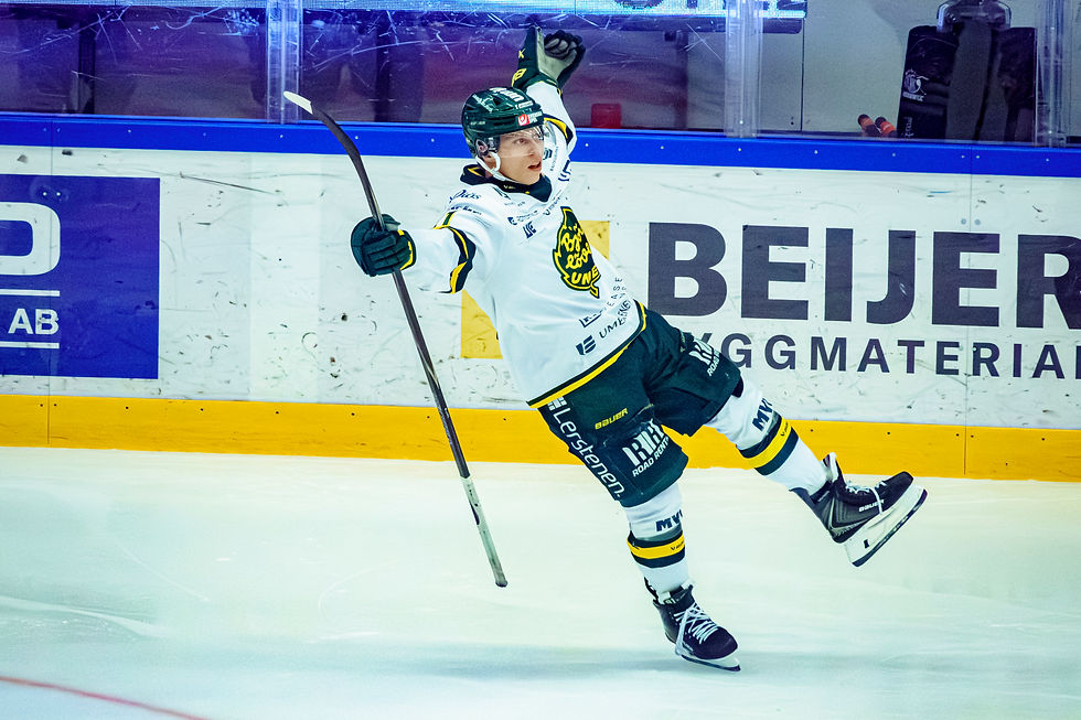 Ishockeyspiller i hvit og grønn drakt jubler med løftet arm på isen. Bakgrunn viser reklamevegger. Energi og glede i bevegelsen.