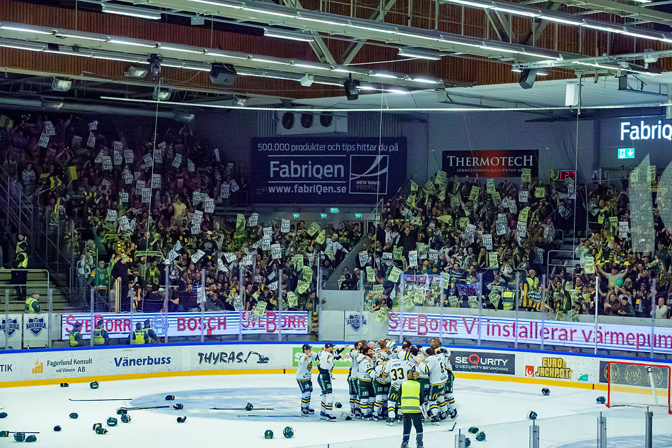 Hockey team huddles on ice with scattered gear. Large cheering crowd in stands holding signs. Arena lit with ads on walls.