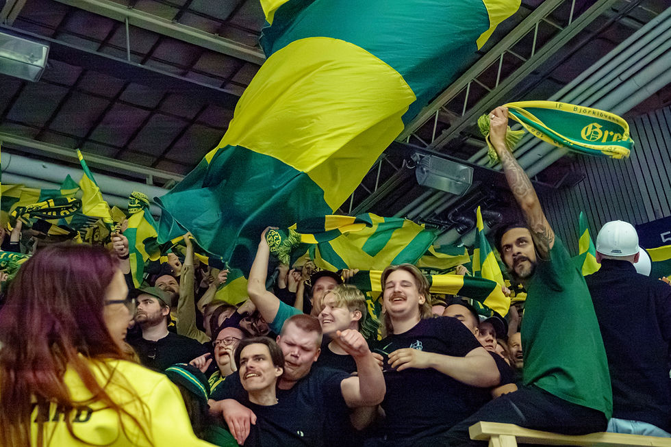 Mennesker heier med gule og grønne flagg i en stadion, uttrykker glede og begeistring. Noen har skjerf med tekst. Energisk stemning.