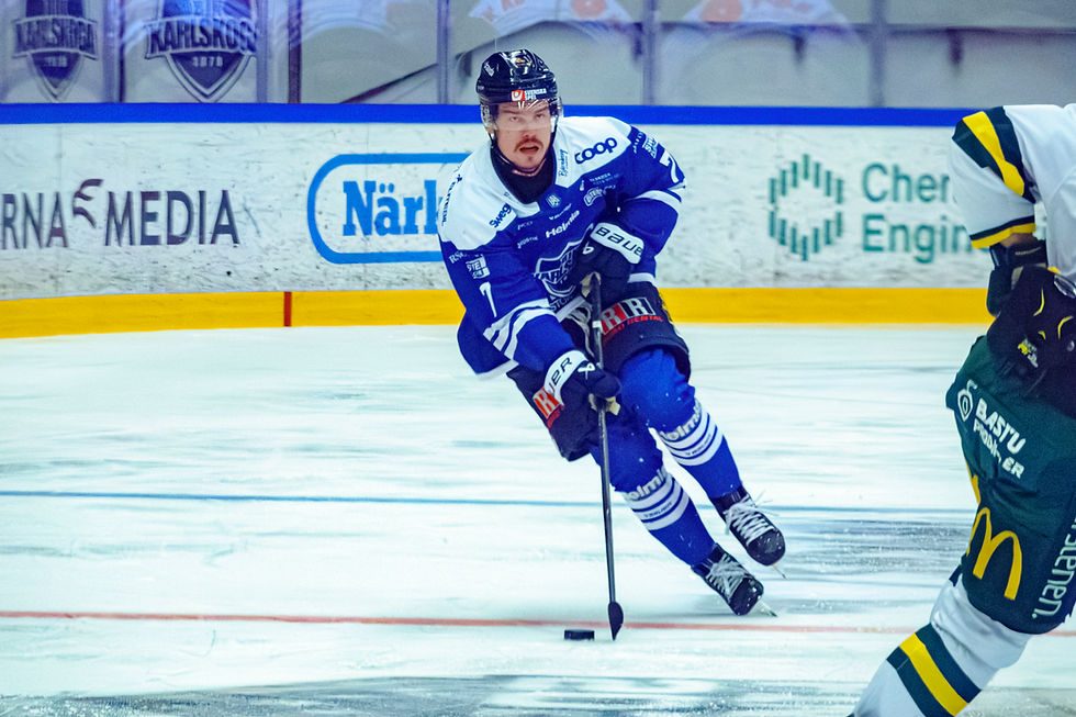 Hockey player in blue uniform skates forward, stick ready, on an indoor rink. Advertisements on boards, teammates nearby, action intense.
