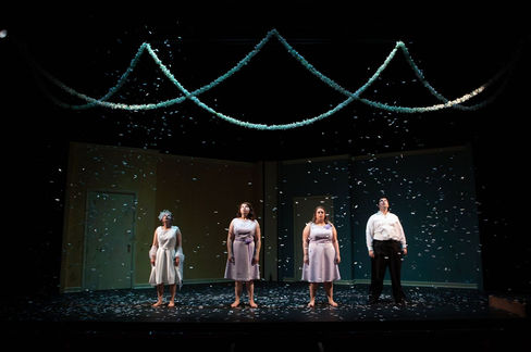 Four actors stand center stage facing the audience. The woman standing furthest stage right is in a tea-length wedding dress with a sheer shawl. Next to her, two women in bridesmaids dresses. None of the women are wearing shoes. Next in the line is a man in black pants and a dress shirt. They are all facing the audience unsmiling as confetti rains down over them.