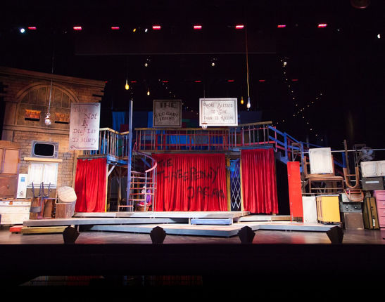 A pre-show shot of the scenery for The Threepenny Opera. It is a large set that appears to have been cobbled together with things picked from the garbage. On stage either side of the stage, a junk wall towers, looking precarious. Center stage, three large red curtains, the center-most with "The Threepenny Opera" spray painted on it in black. Right of center, a metal spiral staircase. Foot lights span the front of the stage.