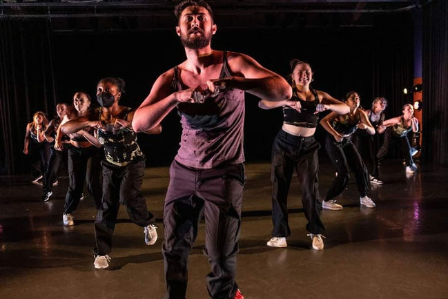Several dancers stand in a "V" formation with their hands in fists at their chest. They appear to be doing sa high energy dance.