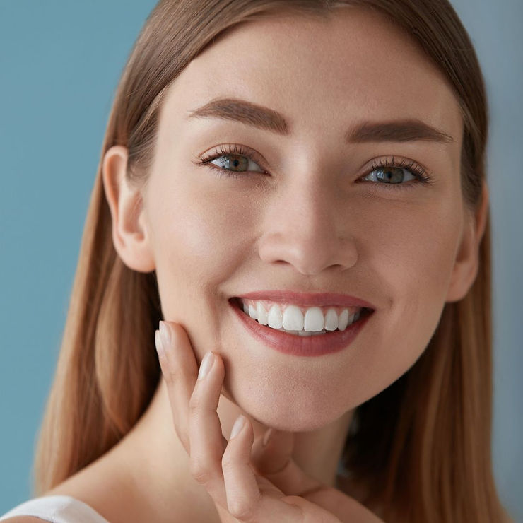 Smiling woman with long hair touches her cheek against a blue background. Her expression is happy and relaxed. No text visible.