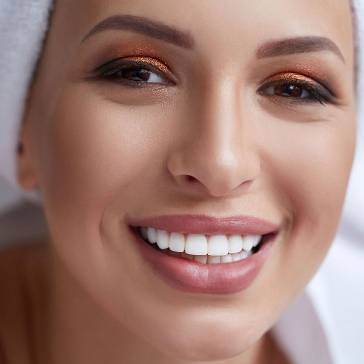 Smiling person with white teeth, wearing a towel on their head. Their eyelids are adorned with shimmering copper eyeshadow. Mood is joyful.