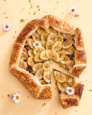 Golden puff pastry galette topped with caramelized banana slices and chocolate nibs, styled with edible daisies on a light yellow background.