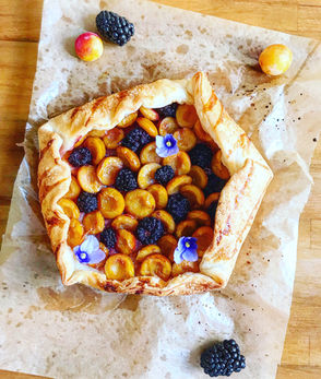 Rustic summer galette filled with golden mirabelle plums and blackberries, topped with edible flowers on brown parchment paper.