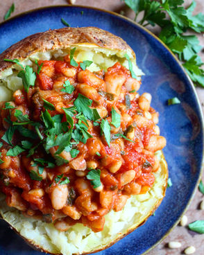 Crispy baked potato split open and generously filled with white beans in a tomato-garlic herb sauce, topped with fresh parsley on a dark blue plate.