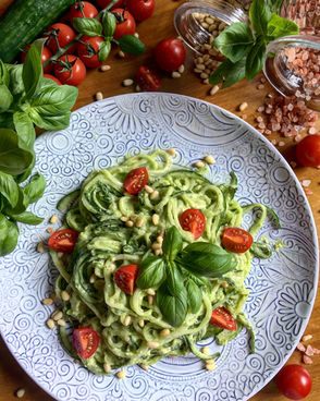 A plate of spiralized raw cucumber noodles tossed in creamy cashew basil sauce, garnished with cherry tomatoes, pine nuts, and fresh basil on a patterned dish.