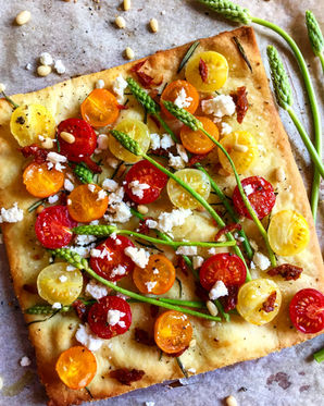 A vibrant summer pizza on a rustic wooden board, topped with sun-dried tomatoes, cherry tomatoes, grilled asparagus, creamy feta, and a drizzle of tomato oil. The crust is golden and crisp, with fresh toppings adding a colorful, nutrient-rich contrast.