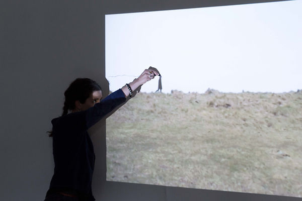 Horizon mobile, 2014 Collaboration avec Juliet Davis Video-projection, performance, dessin Un corps (celui du perfmormeur) traverse la lande. Le plan est fixe, l’horizon légèrement vallonné est centré dans l’image. Le performeur attend devant la projection, son corps, filmé, traverse le champ une première fois. De nouveau, le corps entre dans l’image projetée au mur (toujours de gauche à droite), il fait la taille d’une main. Dans cette mise en abîme, le performeur traverse l’image pour inscrire au mur la traversée du corps, le fusain suivant la tête du personnage. Le corps crée un nouvel horizon à la mesure de sa propre expérience du paysage. La traversée est reproduite, mais réduite à l’échelle d’un paysage, lui-même réduit à l’espace d’exposition. Le dessin qui reste seul au mur fonctionne comme une synthèse et mémoire à la fois d’un geste et de l’expérience du paysage ; il fait le lien de l’image en mouvement vers l’image fixe.