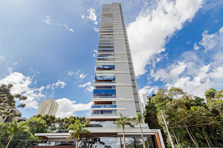 Fotografia profissional de prédio lançamento em Curitiba - Paraná, a foto contém um prédio da base ao topo, o prédio tem amplas vidraças e é cercado de verde, o céu está bem azul.
